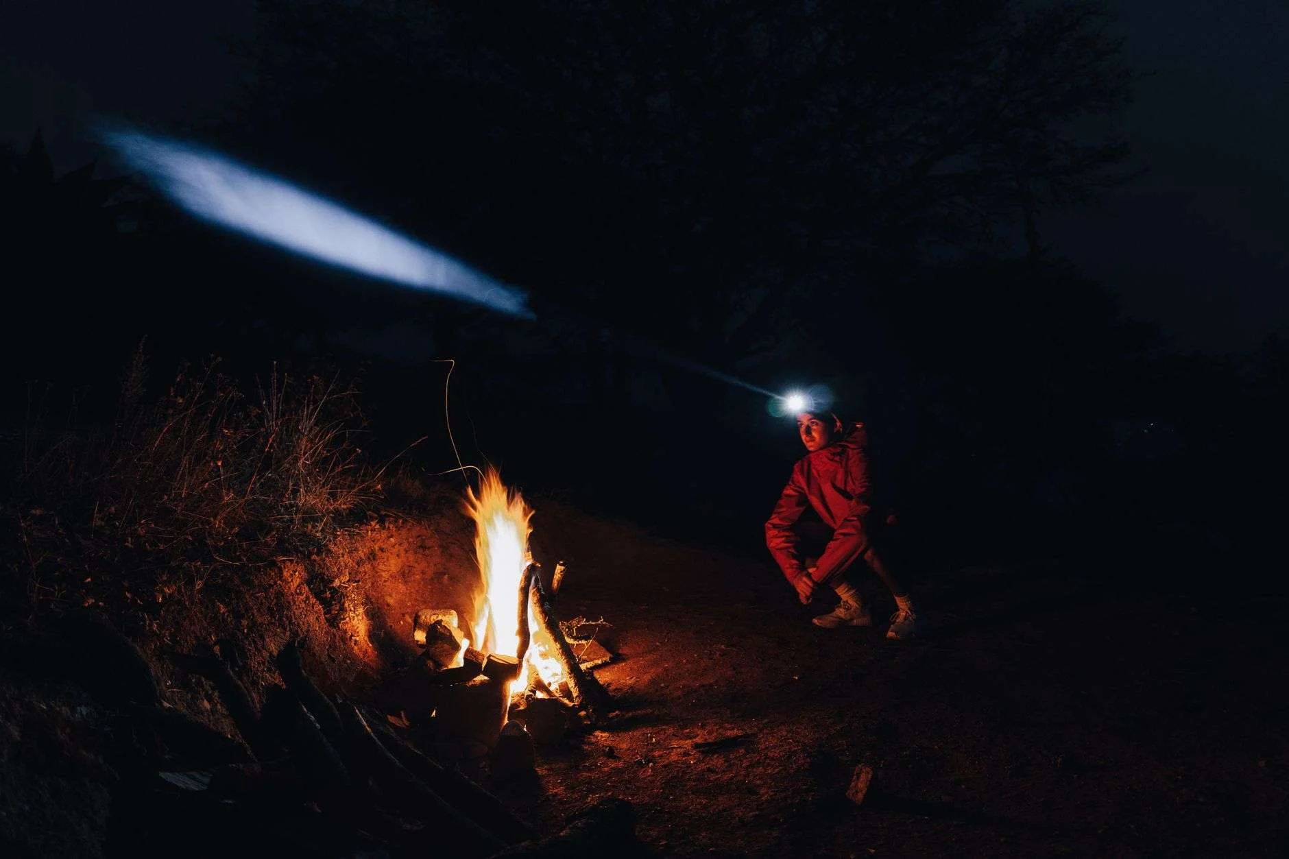 A headlamp beam cutting through darkness beside a camp fire at night