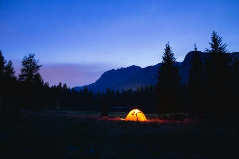 Orange tent lit from inside at dusk below a dark mountain ridge